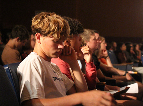 A group of students sit in a darkened auditorium and look at papers.