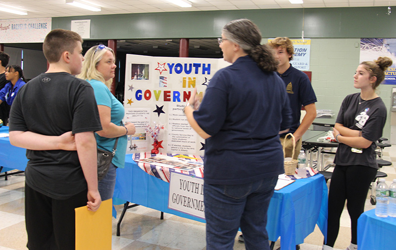 A high school student talks to an adult with dark hair pulled back in a pony tail. They are looking at a poster about Youth in Government. There are others standing around.