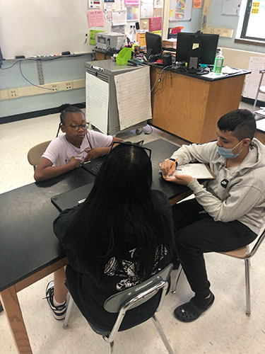 Students sit at tables interviewing others. They have Chromebooks open and are taking notes.