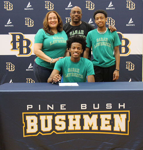 A young man with a green tshirt that says Sarah Lawrence College on it sits at a table with a blue tablecloth that says Pine Bush Bushmen. He is smiling. Behind him are three people - a woman with shoulder-length dark hair, a man with little hair and a young man with short dark hair. They are also wearing green shirts that say Sarah Lawrence except the man in the center whose shirt is black and says Sarah Lawrence.