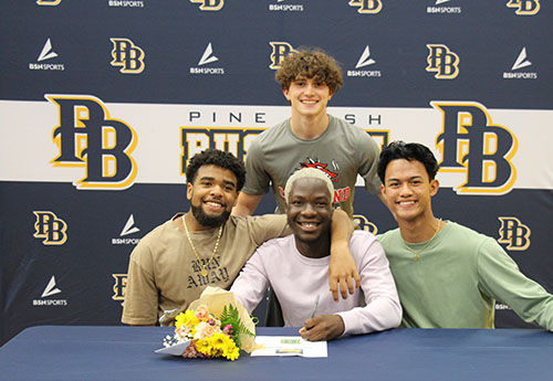 Four young men sit in front of a background that says Pine Bush PB. Three are sitting and one is still behind. The young man on the let has his arm around the one in the center. All are smiling.