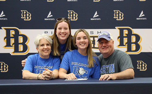 A young woman wearing a blue tshirt that says Barton College sigs at a table. Behind her is a backdrop that says PB and Pine Bush. Next to her are two women and a man. They are all smiling.