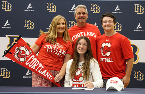 A young woman with long brown hair smiles as she sits at a table signing a document. Standing behind her are a woman with long blonde hair, a man with short gray hair and a young man with short dark hair. All three are wearing red shirts that say Cortland. The woman is holding a red Cortland pennant. Everyone is smiling.