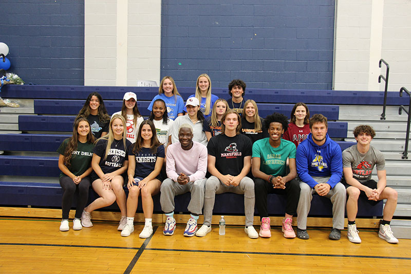 A group of 18 high school students sit in three rows on bleachers. They are all smiling.