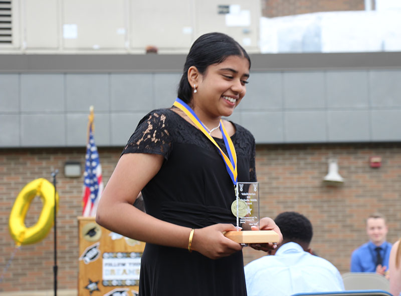 A young woman with long black hair walks back to her seat. She is holding a small plaque and has a medal around her neck. She is smiling.