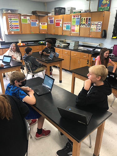 Students sit at tables interviewing others. They have Chromebooks open and are taking notes.