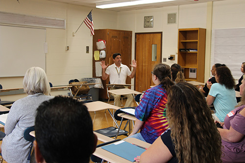 A man in a white polo shirt sits at the front of a class at a desk. He is facing a group of about 15 people also sitting at desks.