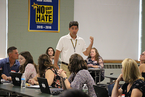 A man in a white shirt and khaki pants stands and talks to a group of adults who are sitting. He has dark hair. There is a No Place for Hate banner in the background.