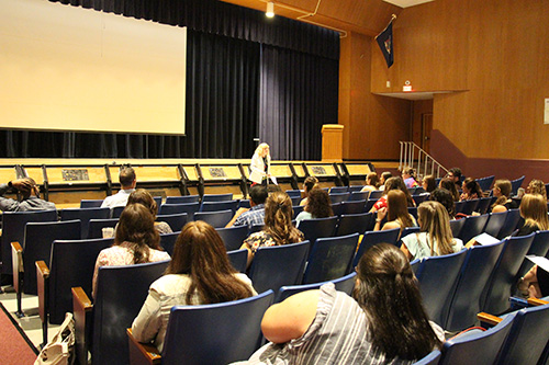 A group of men and women sit in blue seats in an auditorium. There is a woman in front talking to them with blonde hair and yellow jacket.