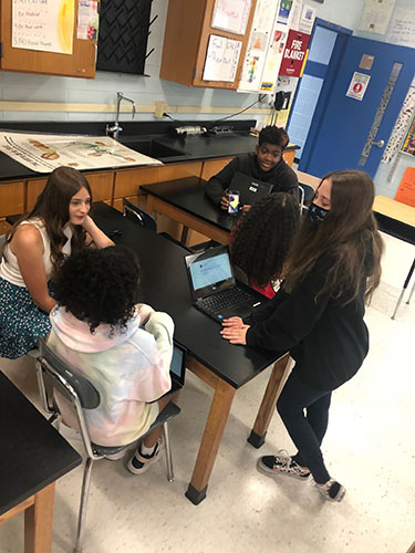 Students sit at tables interviewing others. They have Chromebooks open and are taking notes.