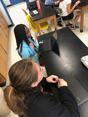 Students sit at tables interviewing others. They have Chromebooks open and are taking notes.