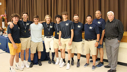 A group of eight high school young men, dressed in navy blue shirts and khaki shorts, stand together, arm in arm, with two adult men on the right, also wearing navy blue shirts. 