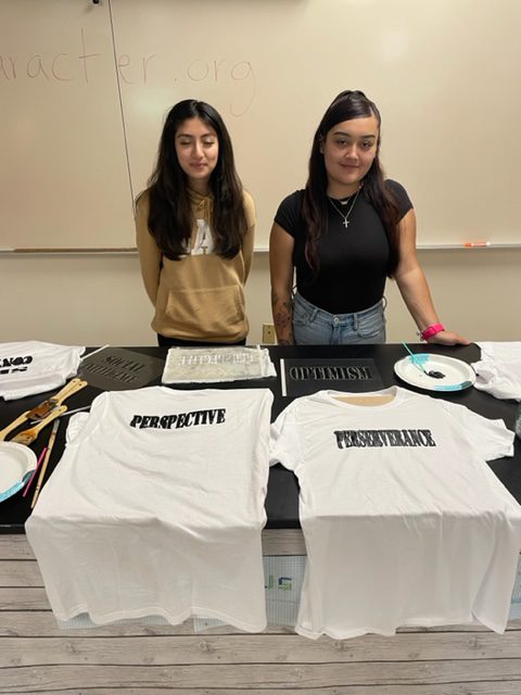 Two high schools girls, both with long dark hair, stand behind a table that has two tshirts on it. One shirt says perseverance and the other perspective.