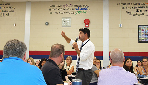 A man wearing a white dress shirt and tie stands amid a group of adults who are listening to him. He is holding a microphone in his left hand and has his right hand in the air.