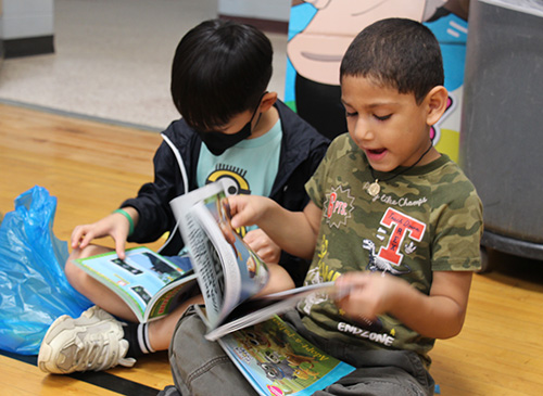 Two elementary age boys sit on a floor and look through the books they just bought. 