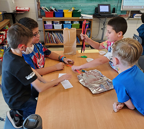 Four elementary age goys sit around a table building with pipe cleaners, aluminum foil and other products.