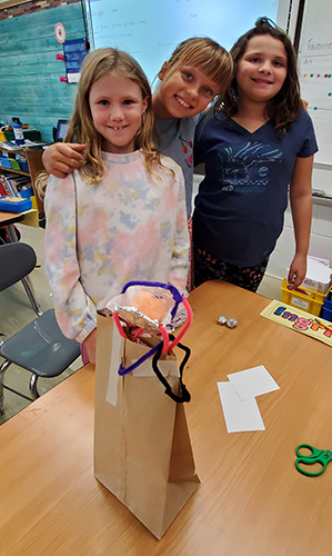 Three elementary students stand and smile with their project in front of them. It is a perch made out of a paper bag, pipe cleaners, index cards and tape.