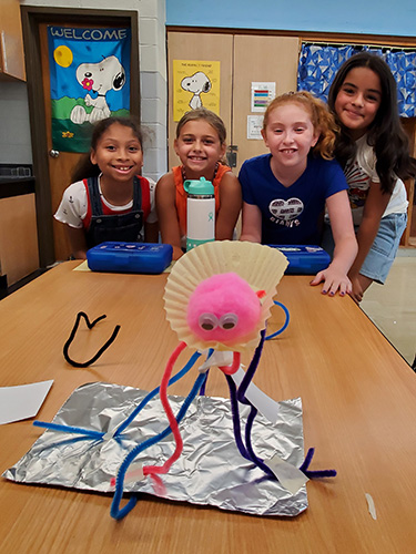 Fou elementary students smile at a table. In front of them is their  project made of pipe cleaners, aluminum foil, tape and index cards. There is a pink pompom sitting in a cupcake paper on top.