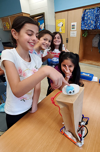 Four elementary age girls smile as they look at their project, made from a paper bag, pipe cleaners, tape and a cupcake paper. Inside the cupcake paper is a blue pompom that one girl is pointing to.