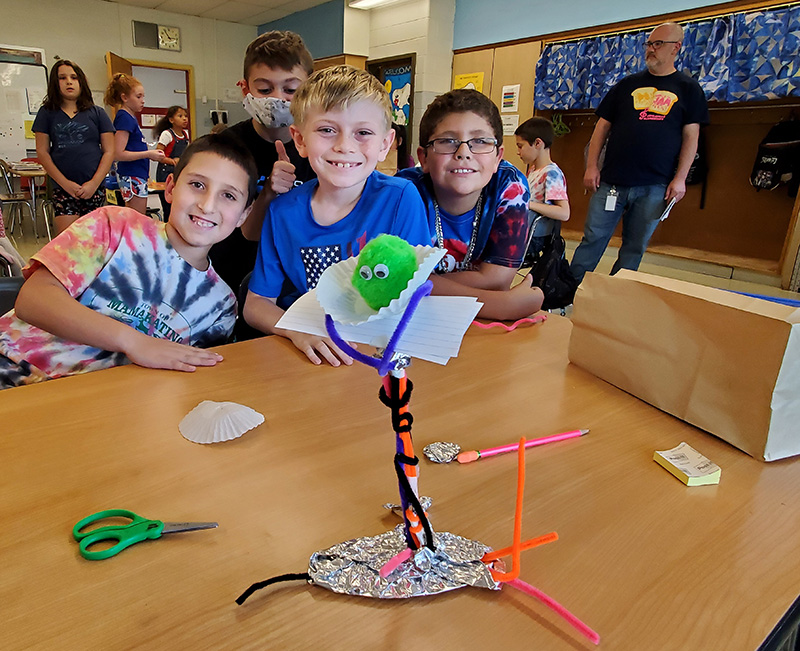 Four elementary boys smile with their project on the table in front of them. They built a perch for a pompom, made out of aluminum foil, pipe cleaners, take, index cards and a cupcake paper.