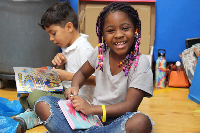 A girl sits cross-legged on the floor. She is opening a book she just bought. She is smiling broadly, wearing a gray shirt and blue jeans, a yellow bracelet and her hair is braided with pink beads in it. There is a boy behind her reading his book.