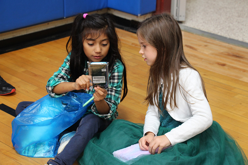 Two elementary age girls sit on the floor looking at a small book the girl on the left bought. She has long dark hair and is wearing a green plaid shirt. She is holding up the book and talking to the girl on the right, who is wearing a green dress and a white sweater.