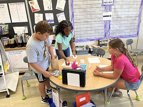 Three fourth-grade students stand around a table, making their project with pipe cleaners, index cards.