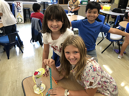 Two fourth-grade girls smile as they look at their project, made from index cards, pipe cleaners and tape. A boy is standing behind them with his arms out smiling.