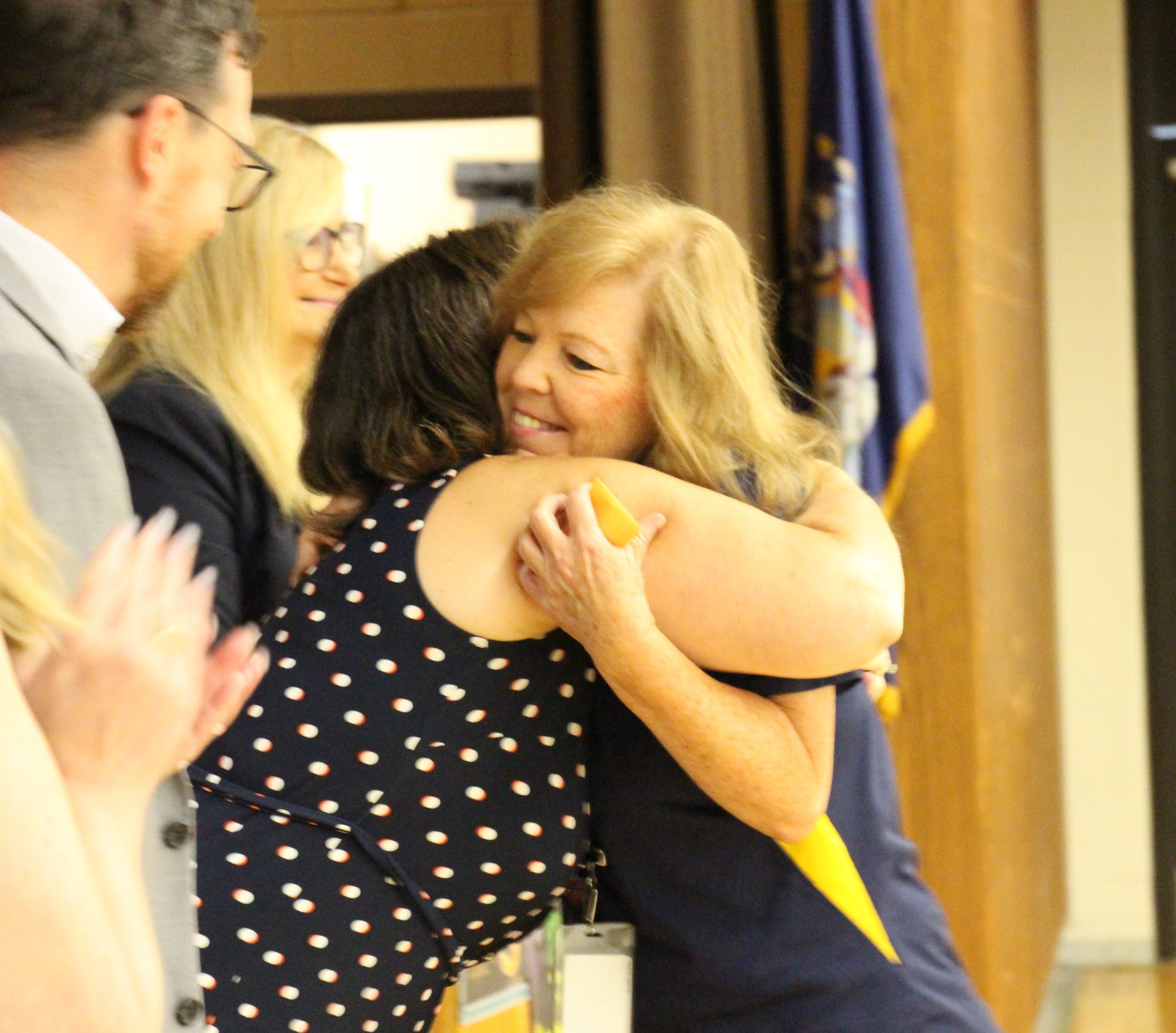 A woman with lighter color hair smiles as she hugs a woman wearing a blue and white polka dot dress.