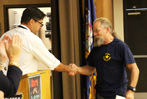 A man in a dark tshirt, with long hair in a ponytail and a beard, shakes hands with a man in a long-sleeve white shirt, dark hair and wearing glasses.