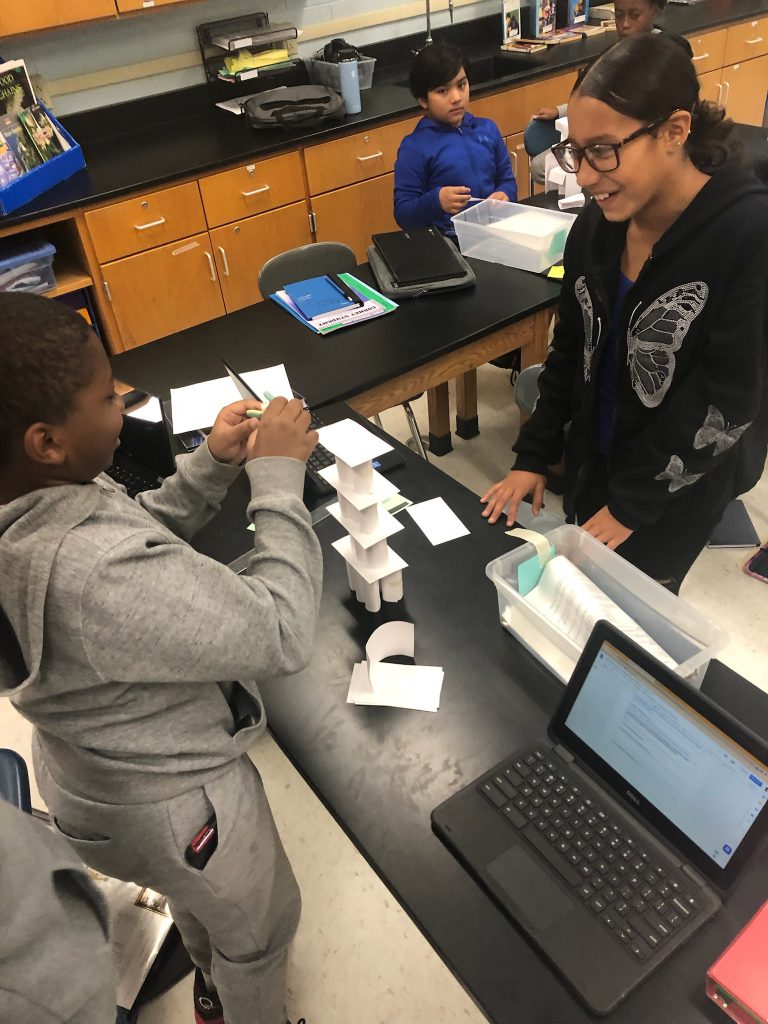 Two middle school age students work on a tower made of index cards in front of them.