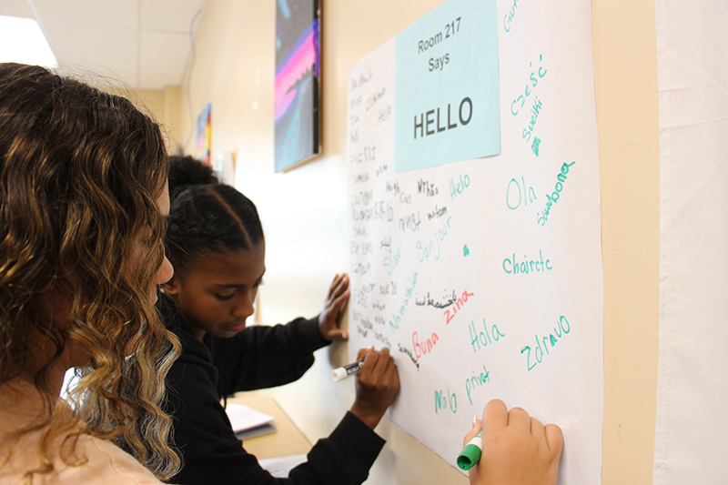 A view from the side - two middle school girls write on a white board many different ways to say hello.
