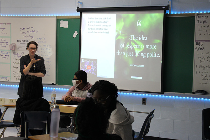 A woman stands at the front of her classroom with students sitting at desks. There is a large screen that says "The idea of respect is more than being polite.".'