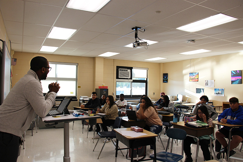 A man at the front of a class leans on a podium as he talks to the class of middle school students. 