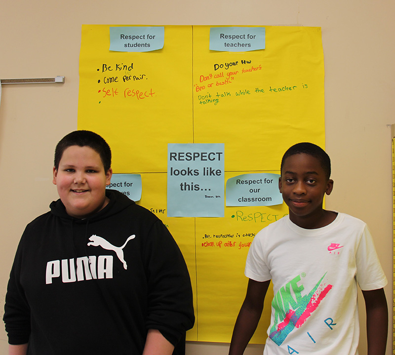 Two middle school boys stand in front of a large yellow poster that has blue papers on it and lots of writing.