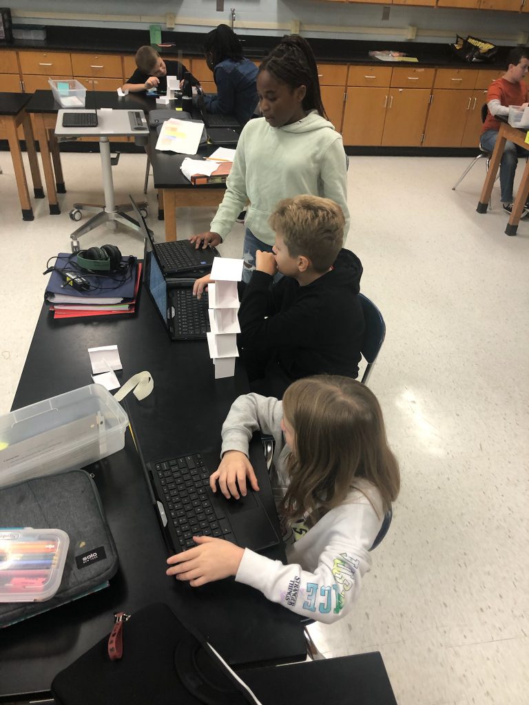 Three middle school students sitting at a black table with a tower made of index cards in front of them.