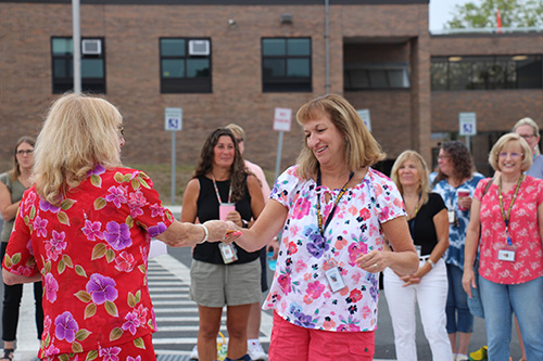 A woman in pink pants and a flowered shirt shakes hands  with another woman in a pink flowered dress who has blonde hair.