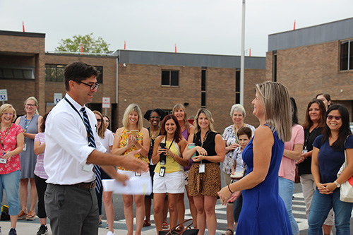 A man in a white dress shirt and tie extends his hand to a woman in a blue dress. She has longer light hair as others around clap. They are outside with a brick building in the background.