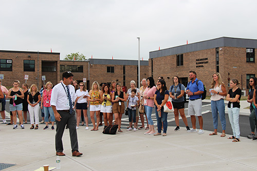 A man in a white dress shirt and tie, with short dark hair, talks to a group of people outside of a brick building.