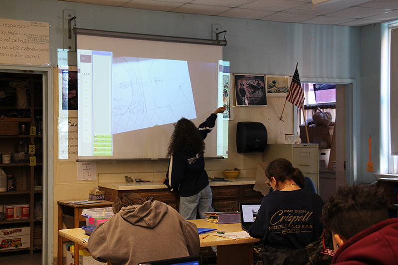 A woman stands at the front of a classroom looking up at a screen that shows dimensions of a face. There are students sitting at their desks watching her.
