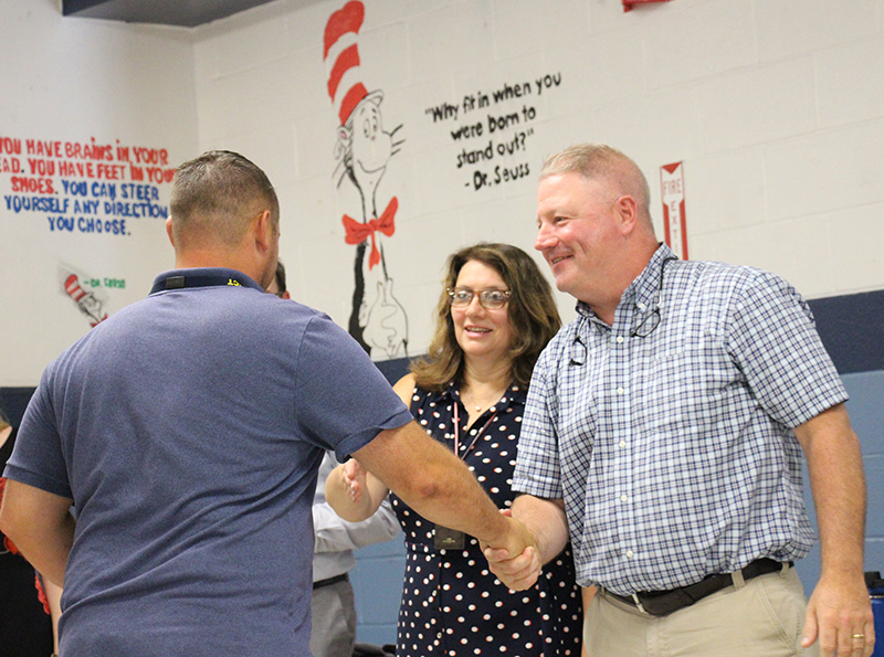 A man in a blue short-sleeve polo shirt shakes hands with a man in a blue plaid dress shirt, who is smiling. A woman with long dark hair, wearing a blue and white polka dot dress stands watching and smiling.