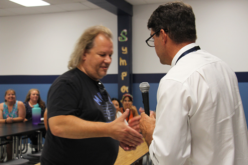 A man with longer blonde hair, wearing a dark tshirt, shakes hands with a man with short dark hair, glasses wearing a white dress shirt.