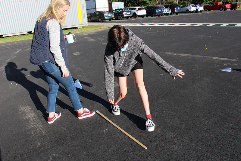 A woman with shoulder-length blonde hair, wearing blue jeans and a long-sleeved shirt and vest, helps a student measure with a yard stick. The student is bending down to pick up the stick along blacktop. The student is wearing black shorts, sneakers and a gray sweatshirt.