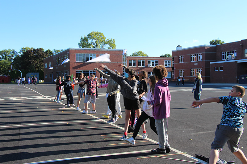 A group of about 20 middle school students stand on a straight line and throw paper airplanes onto blacktop.