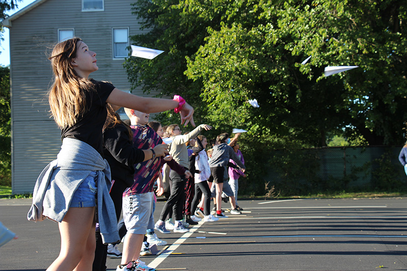A group of about 20 middle school students stand on a white line in a parking lot and throw their paper airplanes forward.