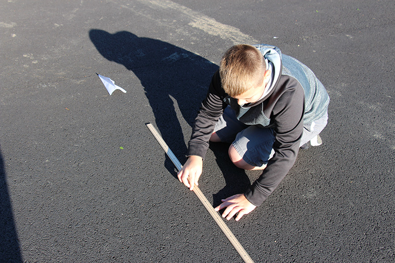 A middle-school boy wearing a denim jacket with black sleeves and hood uses a wooden measuring stick to measure the distance he threw the paper airplane that's on the ground.