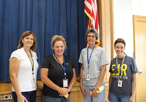 Four women stand in front of a stage with a blue curtain and American flat in the corner. The woman on the left has shorter brown hair and is wearing a white shirt; the woman second from left has her light hair pulled back ad is wearing a dark short-sleeve shirt; the woman second from right has short light hair and is wearing a gray tshirt and the woman on the right has short hair and is wearing a blue shirt with EJR on the front. All are smiling.