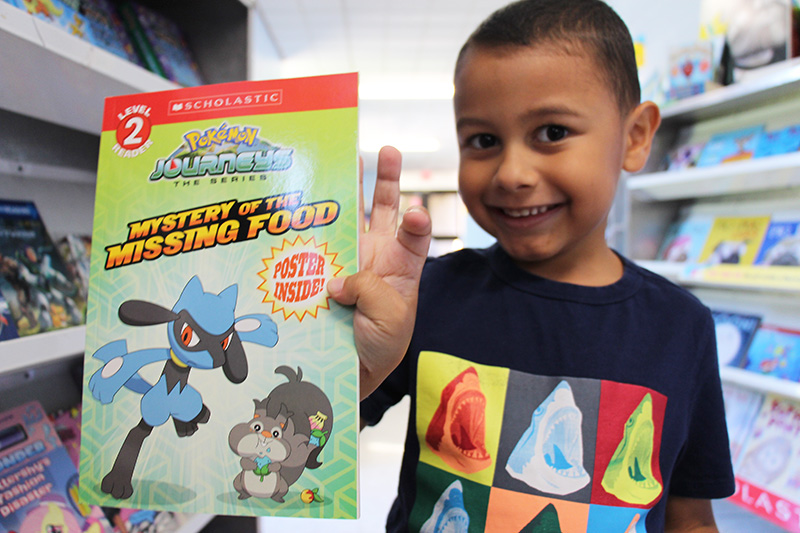 A kindergarten boy with very short dark hair smiles as he holds up a paperback Pokemon book.