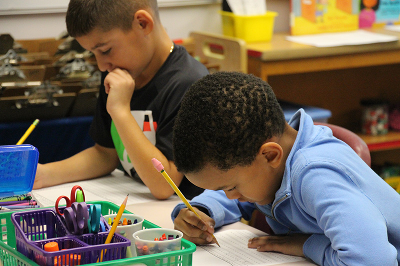 Two second-grade boys use pencils to write on a piece of paper. The boy on the right is wearing a blue shirt and has short dark hair. The boy on the left also has short dark hair and is writing with a pencil as well. There is a green basket on the table in front of them filled with crayons and pencils.