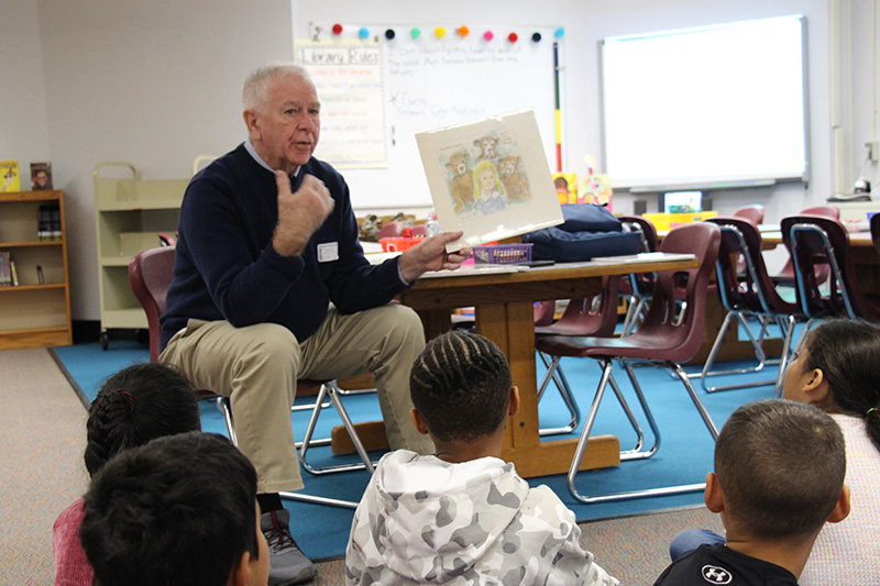 A man with white hair sits and talks to students. He is wearing a blue sweater and tan pants and holding up a drawing of bears. Students sit in front of him watching and listening.